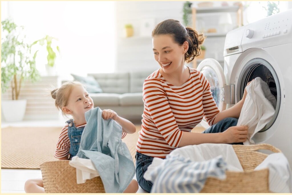 A smiling woman and a young girl, both in striped shirts, are doing laundry together in a sunny living room with a washing machine and a basket of clothes